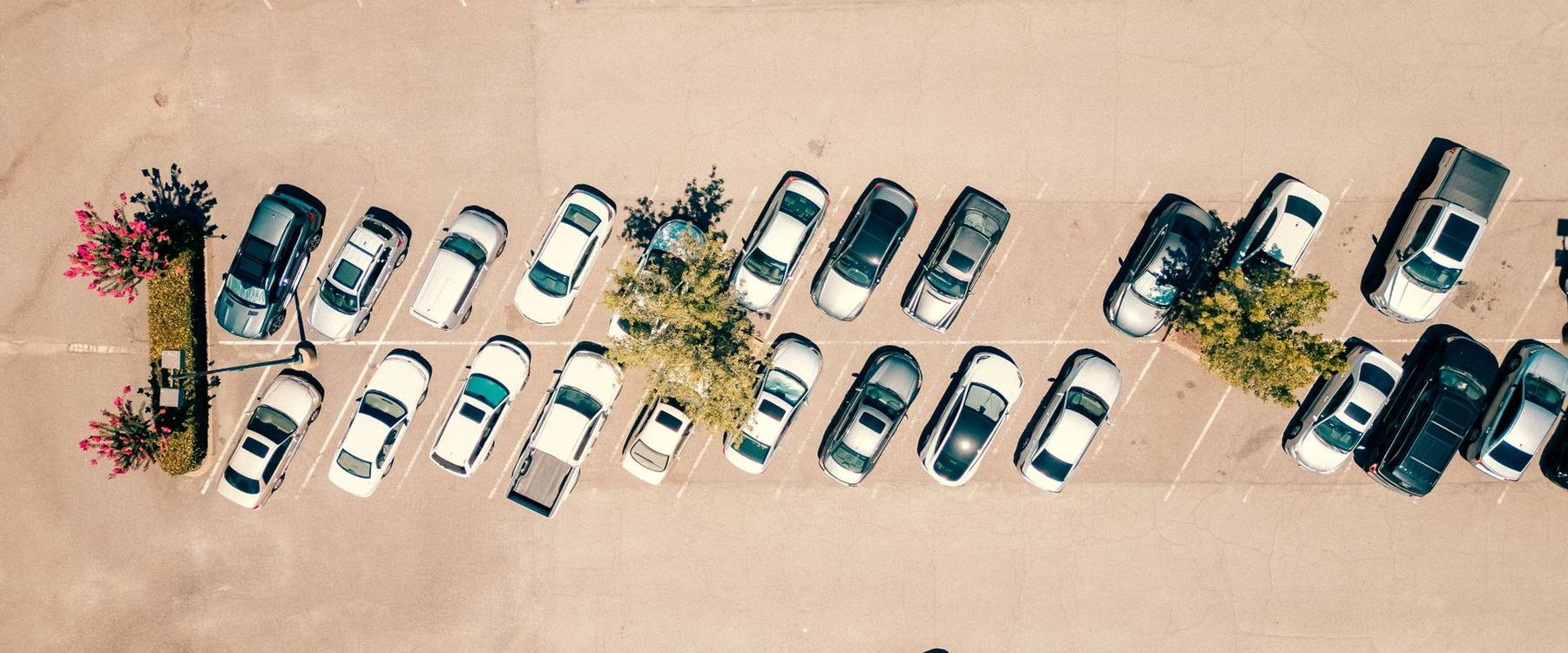Vista aérea de um pátio de estacionamento ao sol, com carros em vagas demarcadas e canteiros floridos